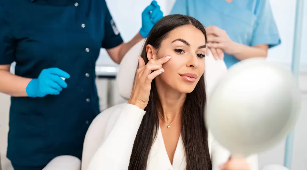 Beautiful and happy young woman sitting in medical chair and looking in the mirror. She is satisfied after successful beauty treatment with hyaluronic acid fillers or botulinum toxin injections.